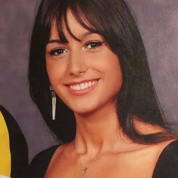 Smiling person with long dark hair and earrings, wearing a dark top against a solid background.